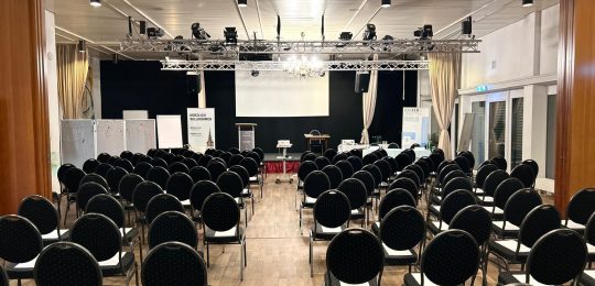 In einem gut beleuchteten Konferenzraum, der für eine Veranstaltung oder ein Seminar eingerichtet ist, stehen Reihen leerer schwarzer Stühle vor einer Bühne mit einem Podium, einer Projektionsfläche und Präsentationstafeln.
