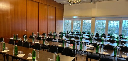 Ein Konferenzraum mit Reihen von Tischen und Stühlen, jeder Tisch ist mit Wasserflaschen, Gläsern und Servietten ausgestattet. Große Fenster geben den Blick auf die Bäume und Gebäude draußen frei. Ein Kronleuchter hängt von der Decke herab.