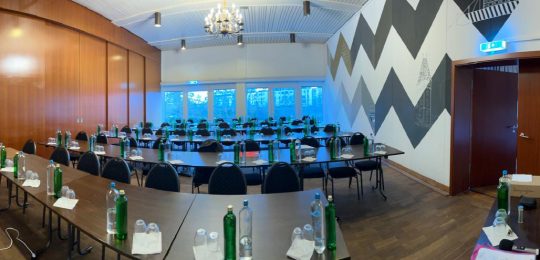 Panoramablick auf einen modernen Konferenzraum mit Holzfußboden, schwarzen Stühlen, langen Tischen mit Wasserflaschen und Notizblöcken, einem Kronleuchter, großen Fenstern, einem Flipchart und geometrischen Wandmustern.