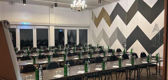 Ein moderner Konferenzraum mit Reihen von Tischen und Stühlen, jeder Platz mit grünen Glasflaschen und Gläsern ausgestattet. Der Raum hat zickzack-gemusterte Wände und einen Kronleuchter, der von der Decke hängt.