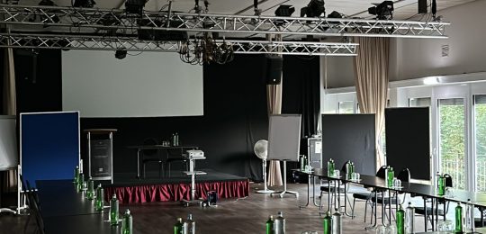 Ein großer Konferenzraum mit schwarzen Stühlen, die um rechteckige Tische angeordnet sind, grünen Flaschen und Gläsern auf den Tischen, Flipcharts und einem Projektor auf der Bühne, mit großen Fenstern, die Tageslicht hereinlassen.