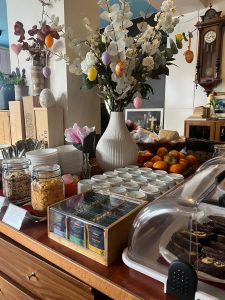 Ein Buffettisch mit Teebeuteln, Müsli, Obst, Gebäck und Gläsern mit Konserven. Weiße Teller und Schüsseln sind fein säuberlich gestapelt, und in der Mitte steht eine Vase mit weißen Blumen und pastellfarbenen Ostereiern.