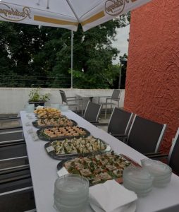 Ein langer Tisch im Freien, bedeckt mit Tellern mit verschiedenem Fingerfood und Vorspeisen, gestapelten Glastellern und Servietten, mit Stühlen drum herum. Im Hintergrund stehen ein weißer Sonnenschirm und Grünzeug.