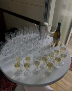 Ein runder Tisch mit zahlreichen Weingläsern, teils mit Weißwein gefüllt, teils leer, daneben eine Weinflasche und ein Metallkrug, neben einer gefliesten Wand und einer Glastür.