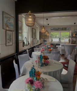 Ein heller, eleganter Speisesaal mit runden, weiß gedeckten Tischen, die mit bunten Blumen und blauen Glasflaschen dekoriert sind. Große Fenster lassen natürliches Licht herein, und von der Decke hängen moderne Pendelleuchten.