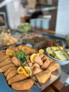 Ein Tablett mit panierten, frittierten und mit Zitronenscheiben und Kräutern garnierten Fingerfoods steht auf einem Buffettisch, während im Hintergrund andere Gerichte und eine Schüssel mit Zitronenscheiben zu sehen sind.
