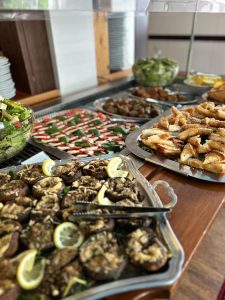 Ein Buffettisch mit Tabletts mit verschiedenen Speisen, darunter gefüllte Champignons mit Zitronenscheiben, frischer Salat, Caprese-Spieße, Fleischbällchen und Gebäck, ordentlich angeordnet mit gestapelten Tellern im Hintergrund.