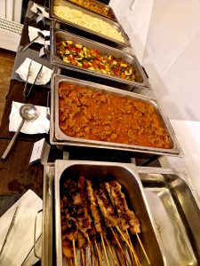 Ein Buffettisch mit Tabletts mit gegrillten Fleischspießen, einem Eintopf mit Soße, gemischtem Gemüse, Nudeln und Reis, dazu Serviergeschirr und Servietten.