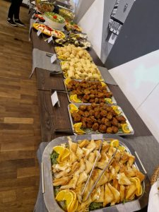 Ein Buffet mit verschiedenen Gerichten, darunter gebratene Vorspeisen, Fleischbällchen, frische Salate und Gebäck, alles auf großen Tabletts mit dekorativen Orangenscheiben auf einem Holzboden angerichtet.