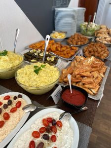 Ein Buffettisch mit Kartoffelsalat, gebratenen Hähnchenflügeln, Fleischbällchen, Fladenbrot, Dips mit Oliven und Tomaten, Zitronenscheiben und gestapelten Tellern im Hintergrund.