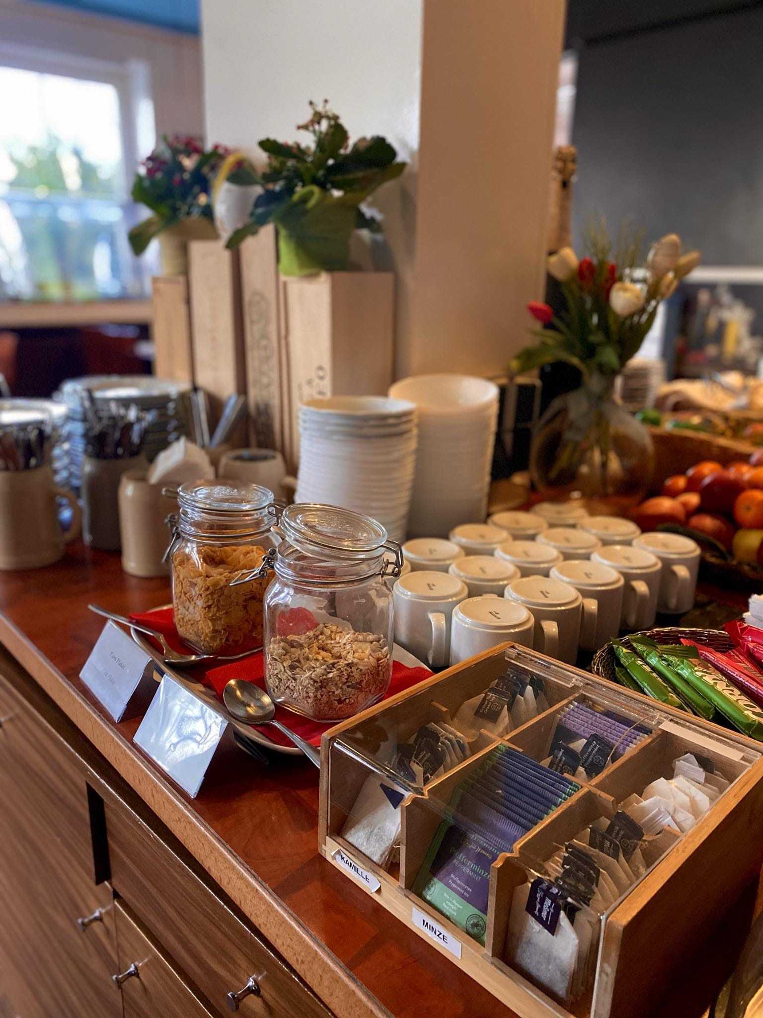 Ein Frühstücksbuffet mit Gläsern mit Müsli, Tassen, Tellern, Besteck, verschiedenen Teebeuteln und einer Schale mit Äpfeln, fein säuberlich auf einem Holztisch mit Blumen im Hintergrund angeordnet.