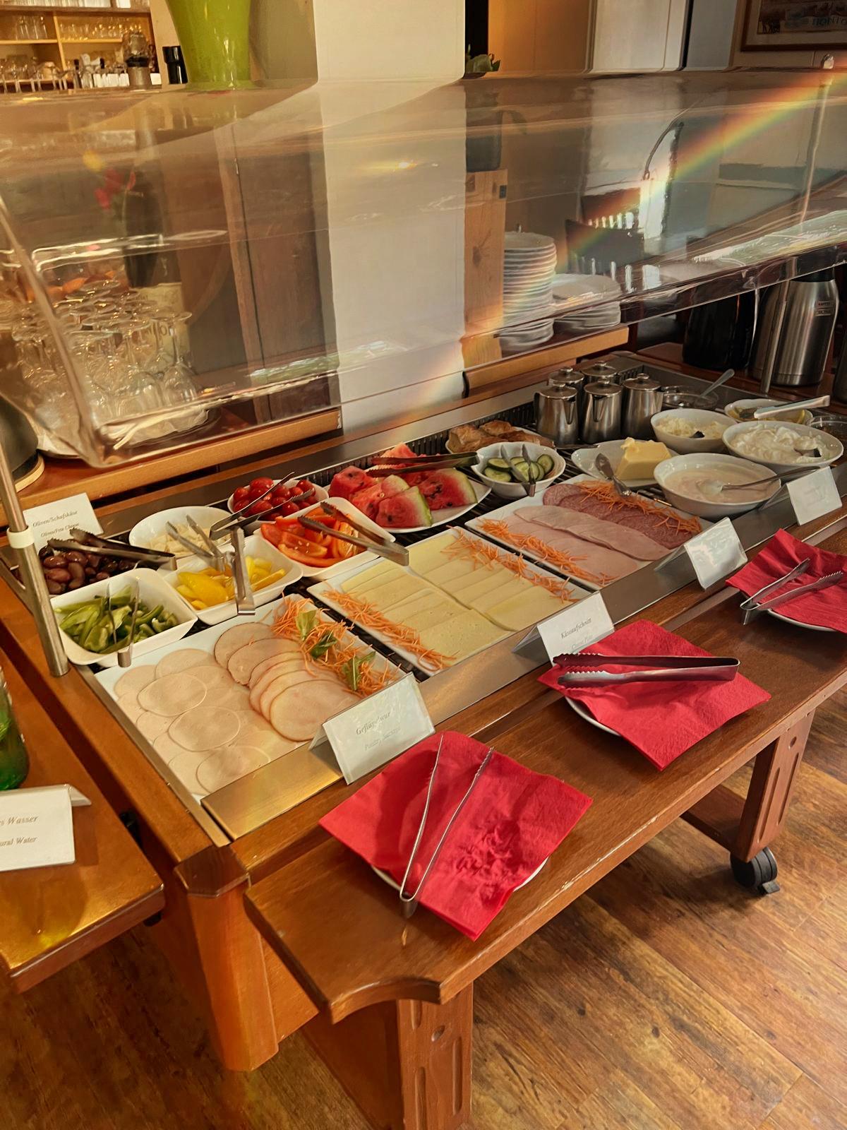 Ein Selbstbedienungsbuffet mit verschiedenen Wurst- und Käsesorten, Gemüse, Essiggurken, Aufstrichen und frischem Obst auf einem Holztisch mit roten Servietten und einem durchsichtigen Hustenschutz.