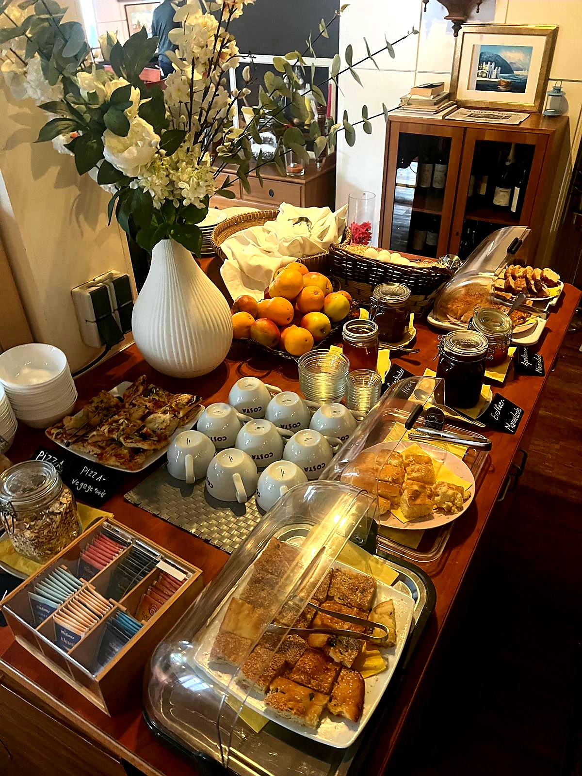 Ein Holztisch ist mit Frühstücksartikeln gedeckt, darunter Gebäck, Brot, Marmelade, Teebeutel, Orangen, Tassen und Teller. Eine weiße Vase mit Blumen schmückt den Tisch, und Körbe enthalten weitere Lebensmittel.